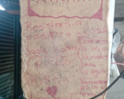 In the image there is a board that has written with red color "Xinka" and says "peaceful resistance" "xinka people" and a drawing of heart. In the background there are some tables and sheets.